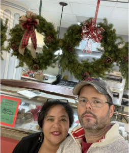 Peace Mitchell (featured left) and Michael Birchard (featured right). Peace is wearing glasses on her head and red lipstick and is smiling at the camera. Micheal is wearing glasses and a hat and looking at the camera.